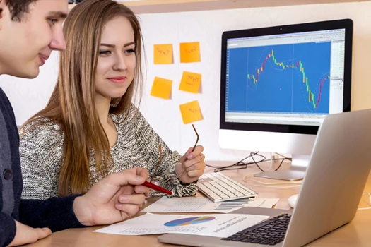 Two people sitting in front of a Laptop with a screen in the backgrond that shows a graph